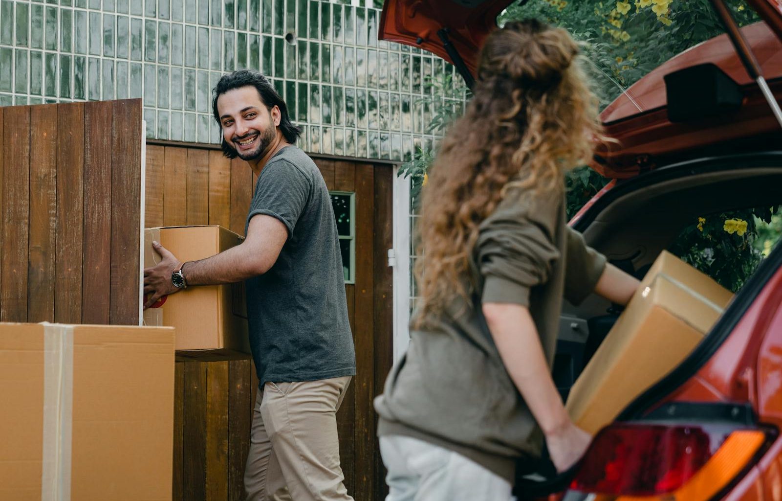 Cheerful modern male and female in comfortable casual clothes unpacking car trunk in green garden of new house while moving in together