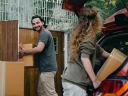 Cheerful modern male and female in comfortable casual clothes unpacking car trunk in green garden of new house while moving in together