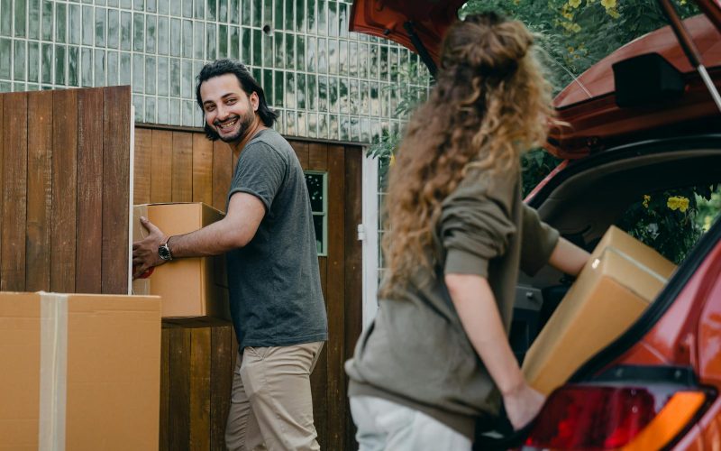 Cheerful modern male and female in comfortable casual clothes unpacking car trunk in green garden of new house while moving in together