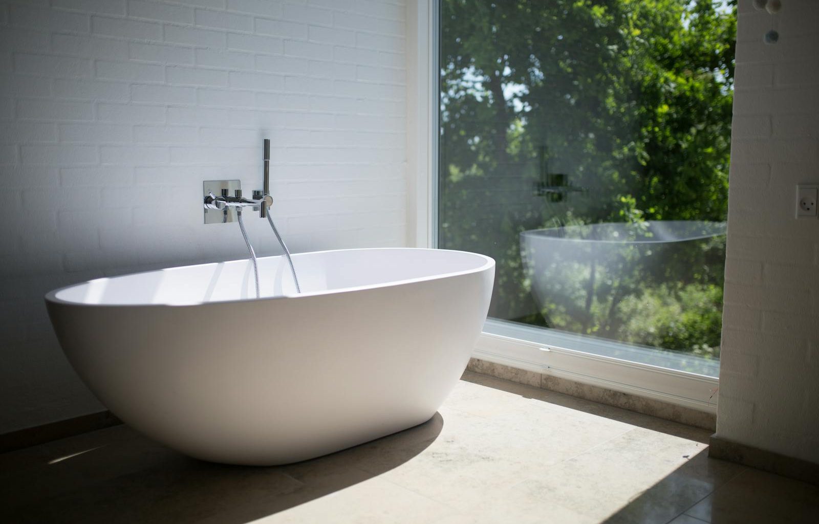 Sleek white bathtub by large window in sunlit minimalist bathroom setting.