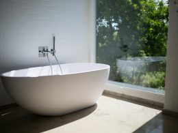 Sleek white bathtub by large window in sunlit minimalist bathroom setting.