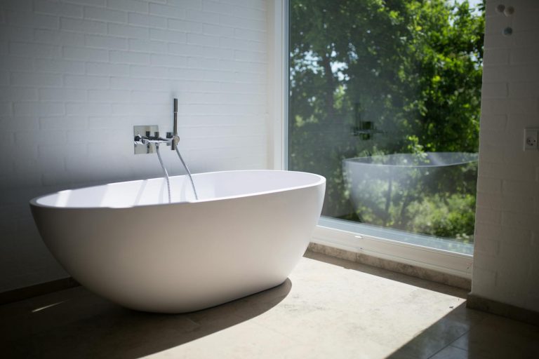 Sleek white bathtub by large window in sunlit minimalist bathroom setting.