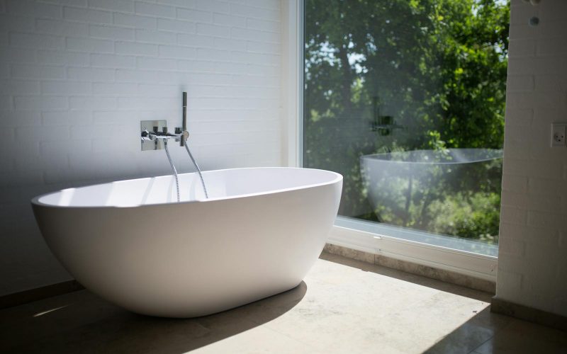 Sleek white bathtub by large window in sunlit minimalist bathroom setting.